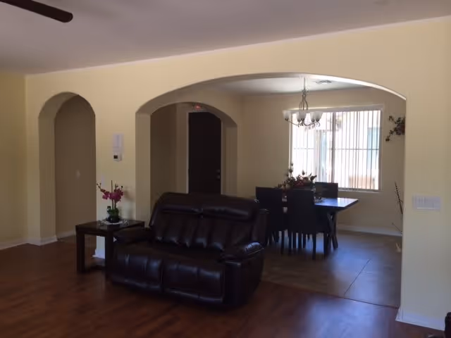 Interior view of a living and dining area in a senior living facility. The foreground features a dark brown leather loveseat on a wooden floor. To the left, there is a small side table with a potted plant and flowers. The background shows a dining table with six black chairs under a chandelier, next to a window with vertical blinds. The walls are painted light yellow, and there are arched doorways leading to other rooms.