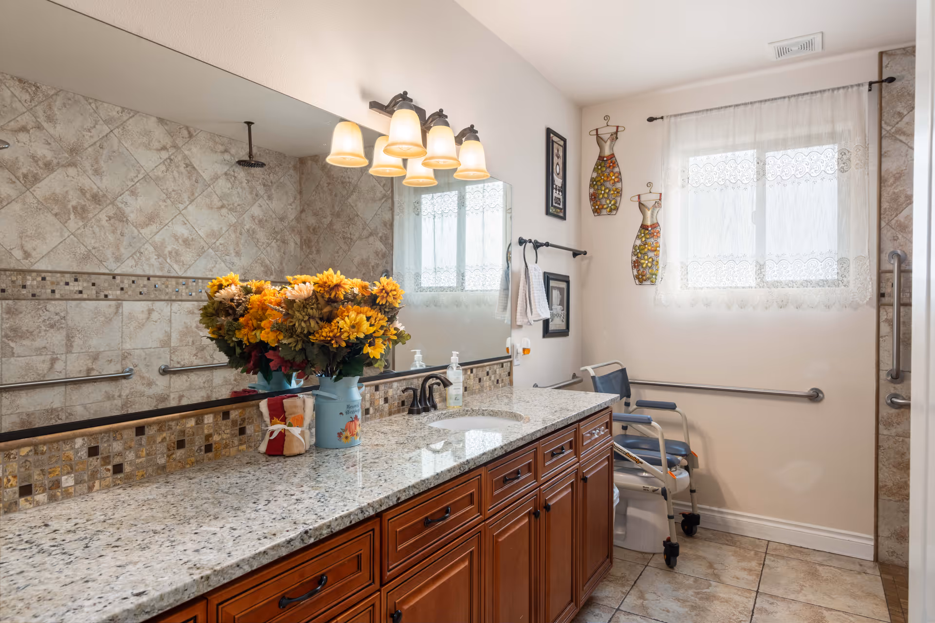 A bathroom with a large granite countertop and wooden cabinets. There is a bouquet of yellow and orange flowers in a blue vase on the counter. The wall behind the counter has a large mirror with four light fixtures above it. The walls are tiled with beige and brown tiles, and there is a window with lace curtains letting in natural light. A shower with a rain showerhead and grab bars is visible, along with a shower chair and additional grab bars on the walls for accessibility.