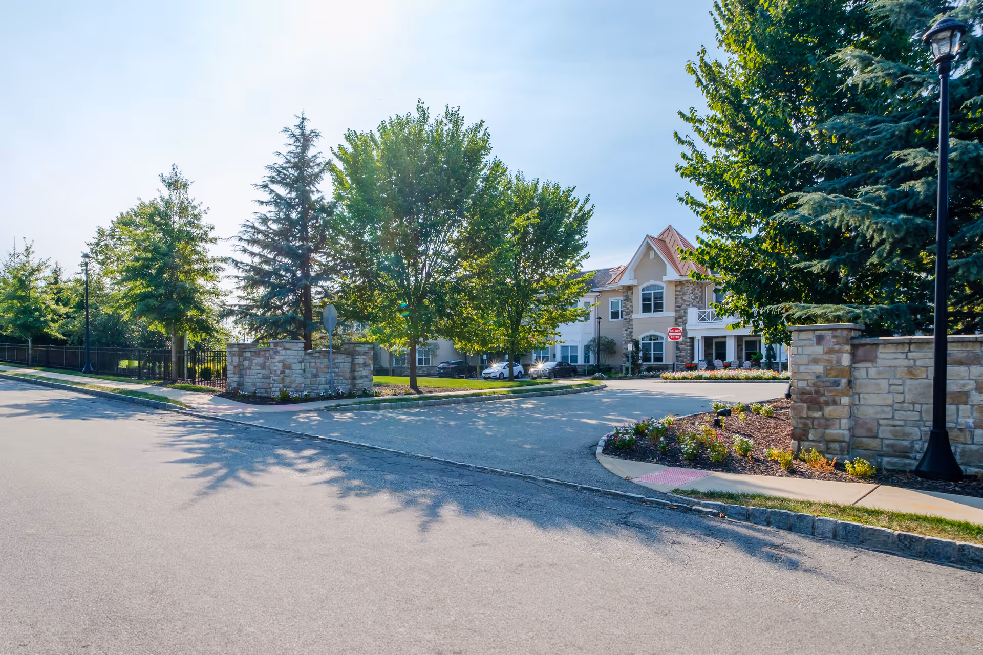 Exterior view of Brandywine Livingston by Monarch senior living facility showing a paved driveway, landscaped garden beds with flowers, stone walls, tall trees, and a two-story building with a red roof and white trim in the background under a clear blue sky.