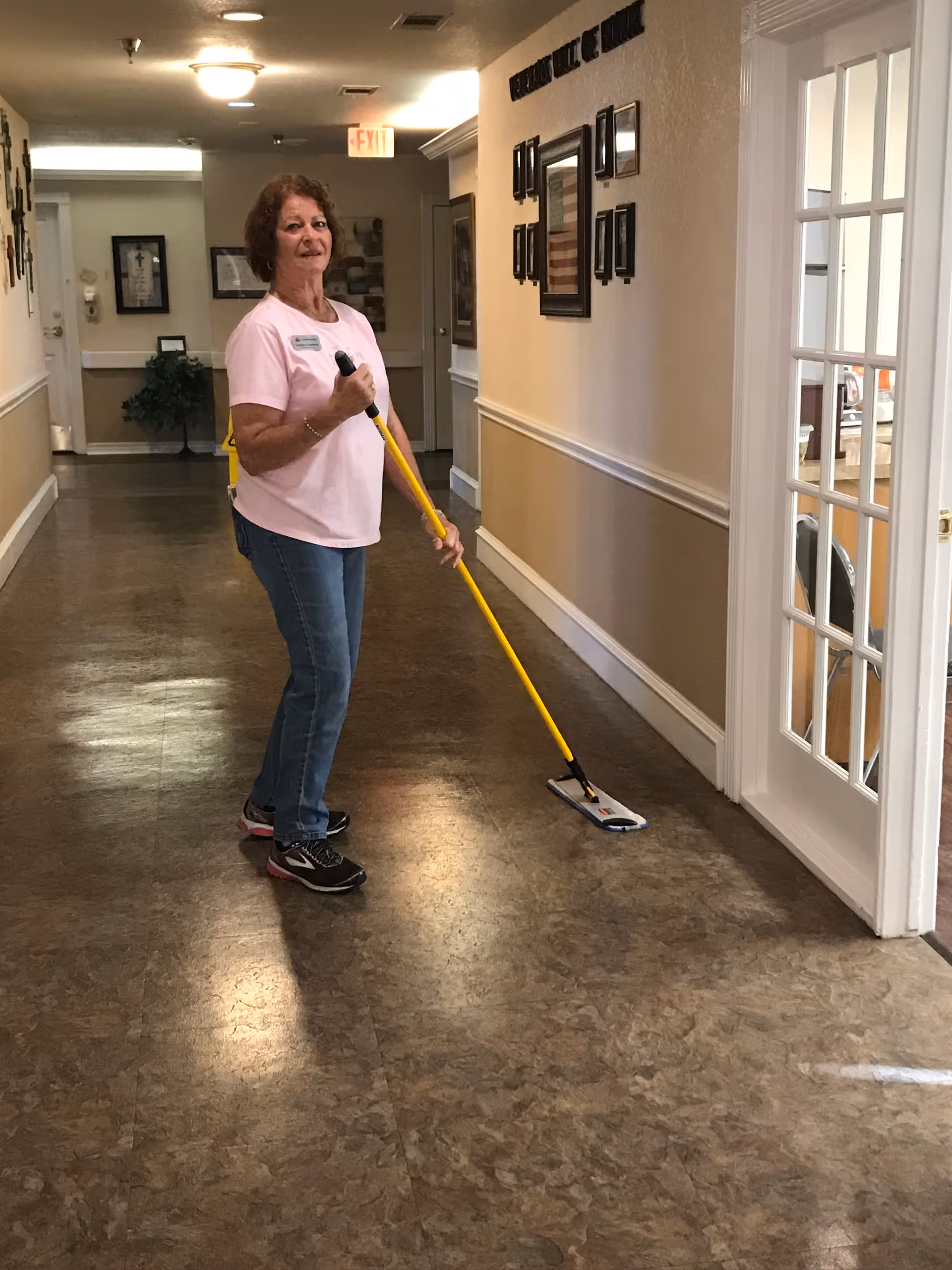 A woman wearing a pink shirt and jeans is mopping the floor in a hallway of a senior living facility. The hallway has beige walls with white trim, framed pictures on the wall, and a glass door on the right side. There is an exit sign visible at the end of the hallway.