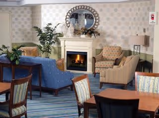 A cozy living room area with a fireplace, a round decorative mirror above it, and various upholstered chairs and sofas arranged around the room. There is a tall plant in the corner, a floor lamp, and a table with chairs in the foreground. The room has patterned wallpaper and a striped carpet.
