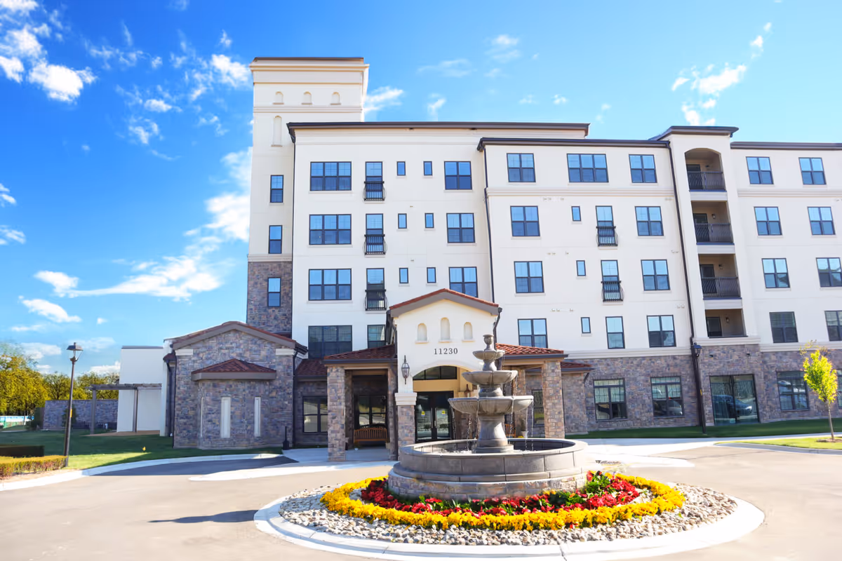 Front exterior view of a multi-story senior living facility building with a stone and stucco facade, a central entrance with the number 11230 above it, and a tiered water fountain surrounded by colorful flowers in the circular driveway in front.