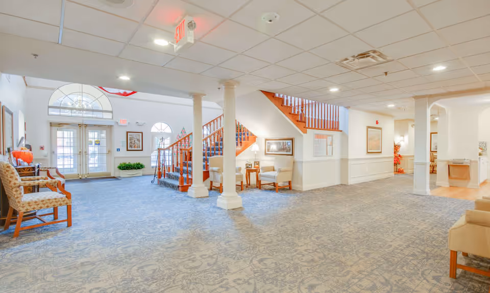 A spacious and well-lit senior living facility lobby with patterned blue carpet, wooden staircase with carpeted steps, several chairs and small tables, framed artwork on the walls, and double glass entrance doors with arched windows above them.