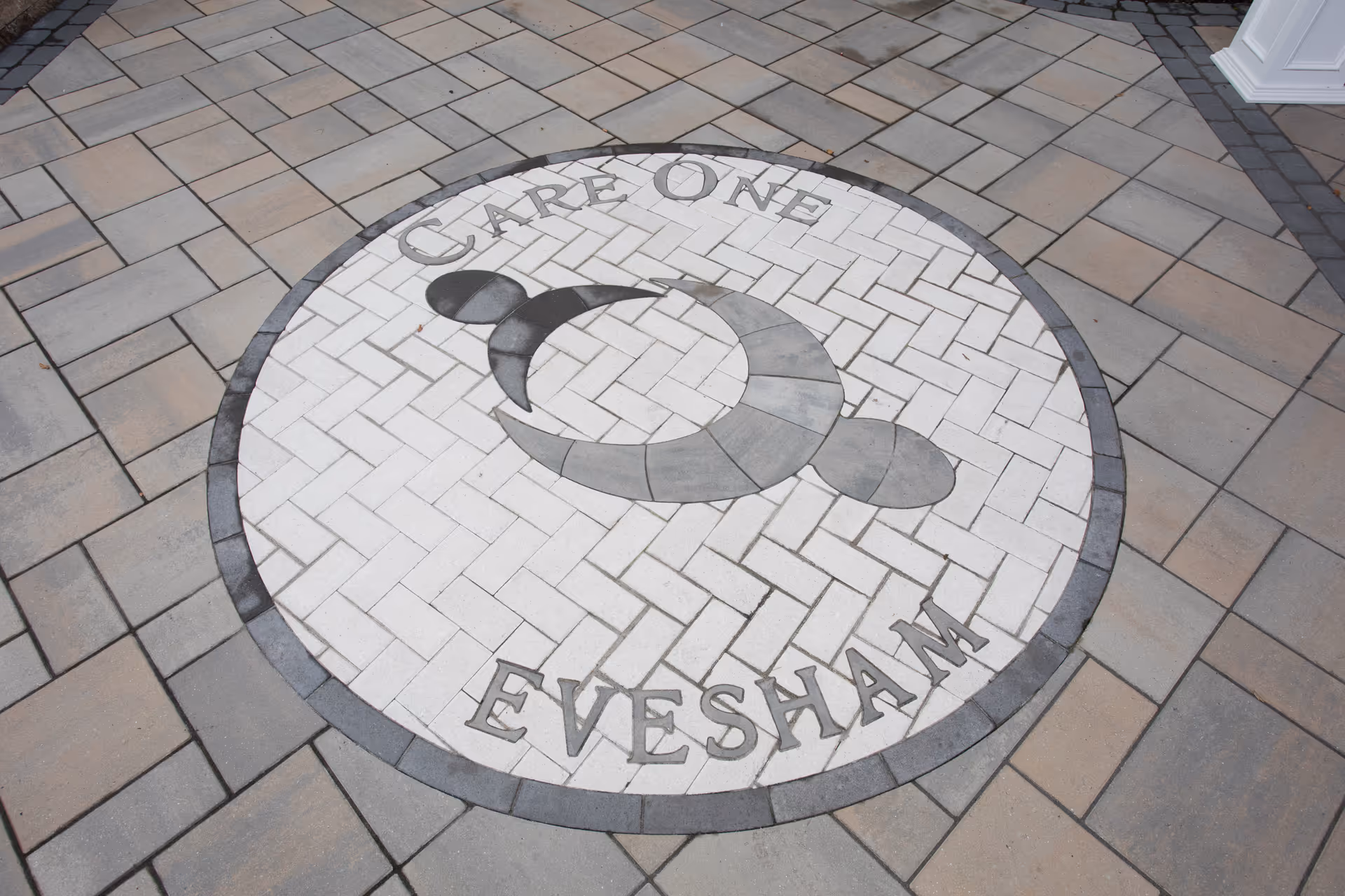 Circular tiled emblem on the ground with the text 'Care One Evesham' surrounding a stylized human figure logo, set within a paved area.