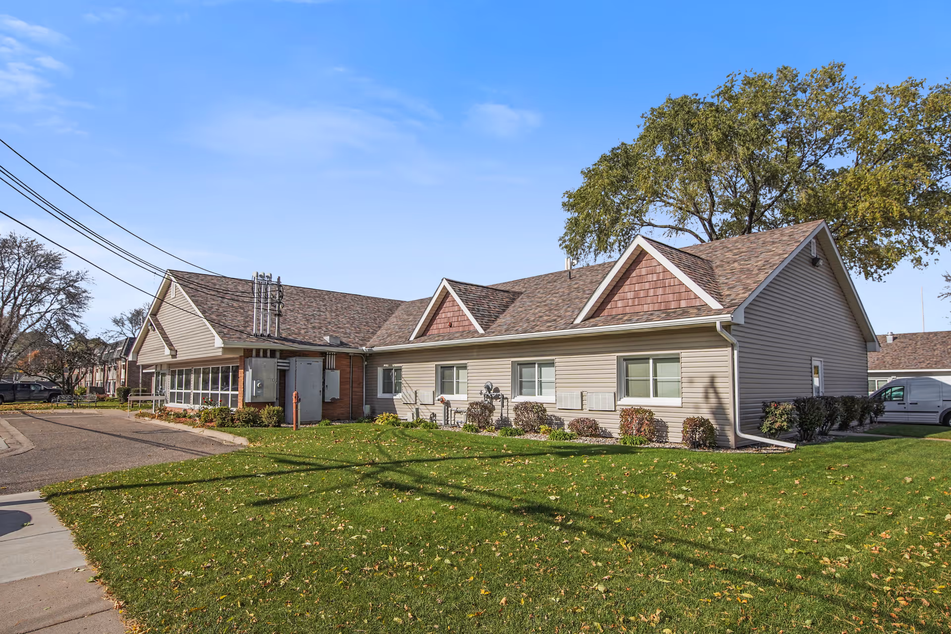 Exterior view of a single-story building with beige siding and brown shingled roof, surrounded by green grass and some bushes. There are power lines overhead and a clear blue sky with a few clouds.