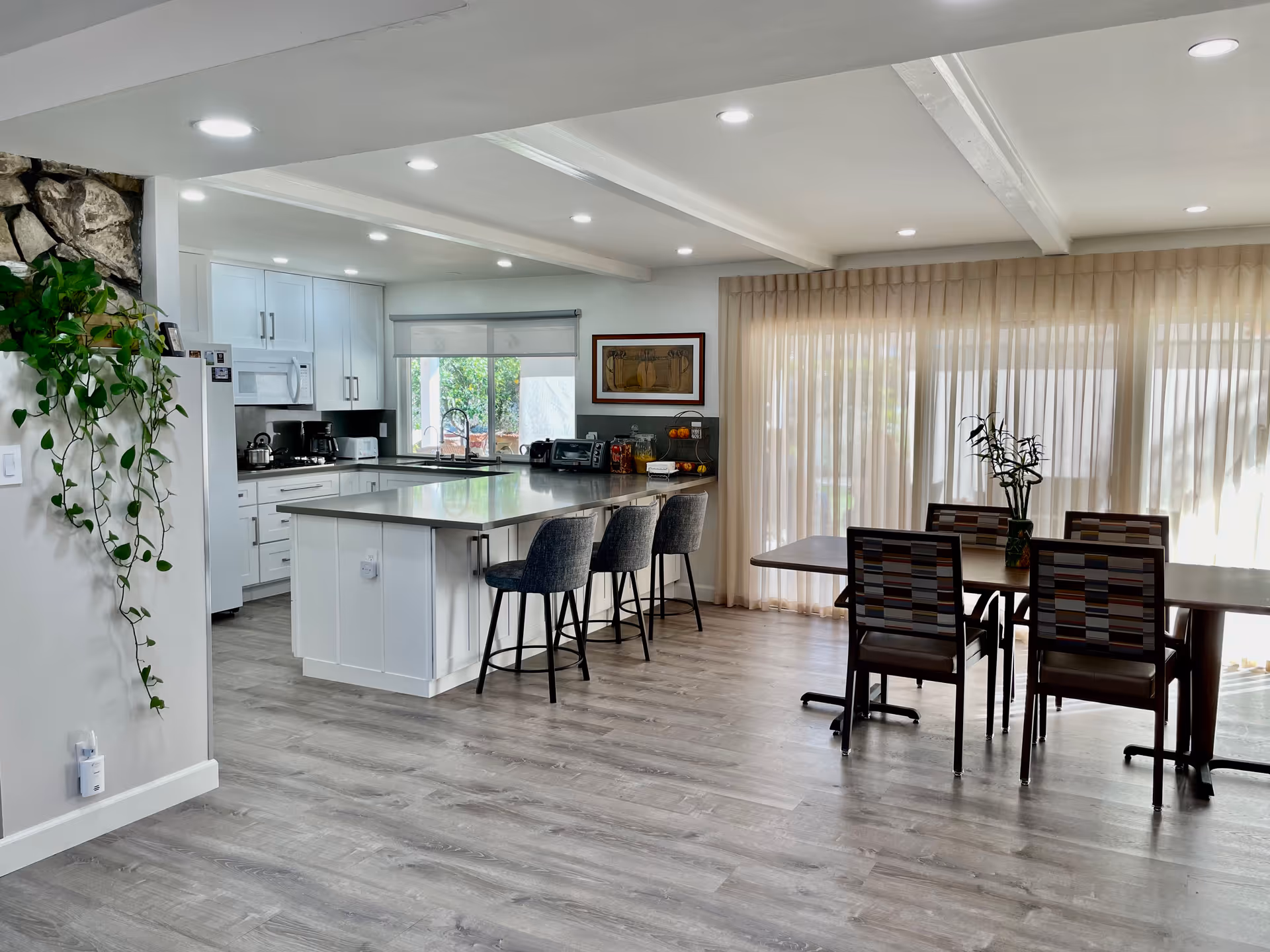 Bright open-plan kitchen and dining area with a central island, bar stools, and a dining table by sheer curtained windows.