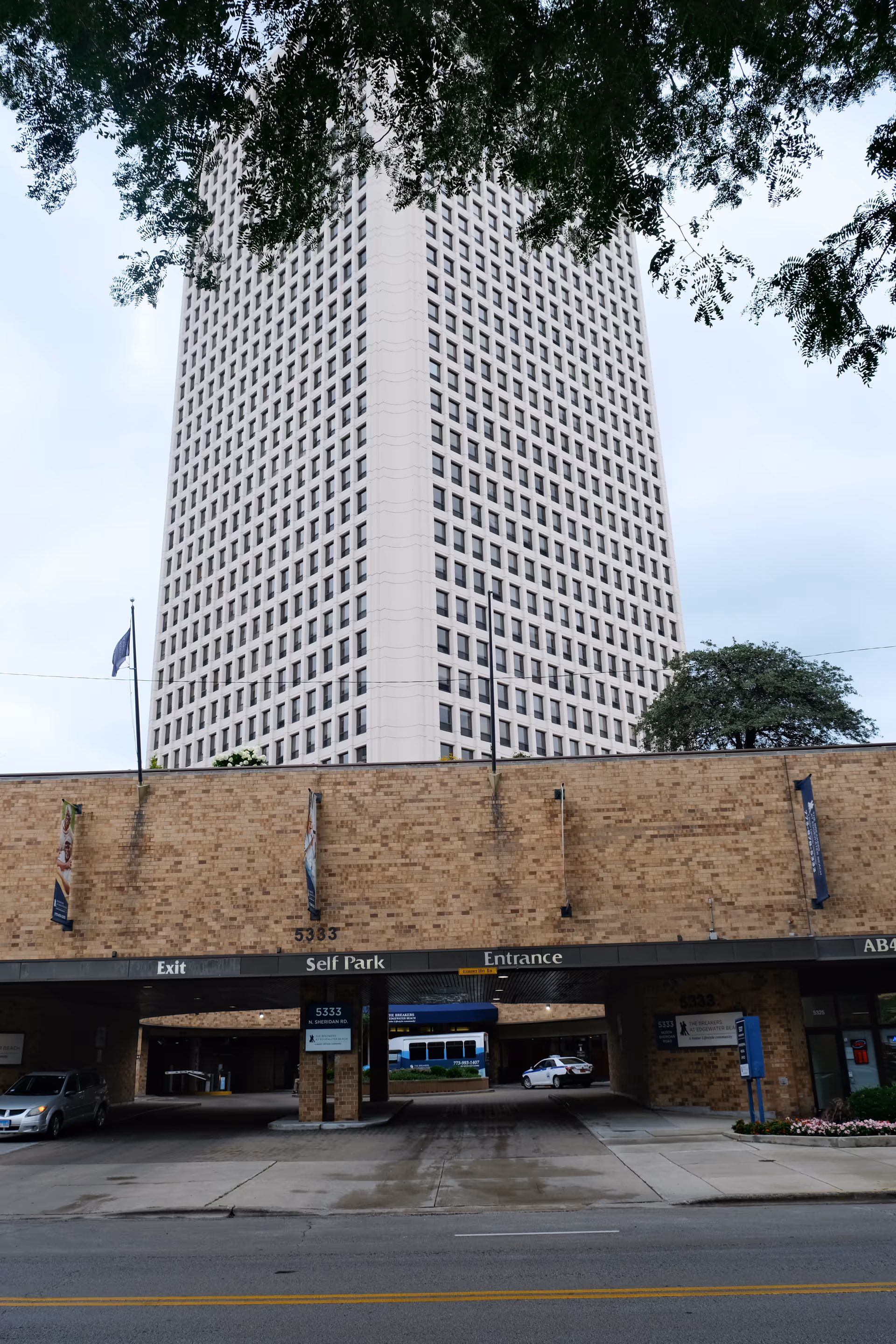 Exterior view of a tall building with a brick parking garage entrance below. The entrance has signs indicating Exit, Self Park, and Entrance. There are a few cars parked near the entrance, and trees partially frame the top of the image.