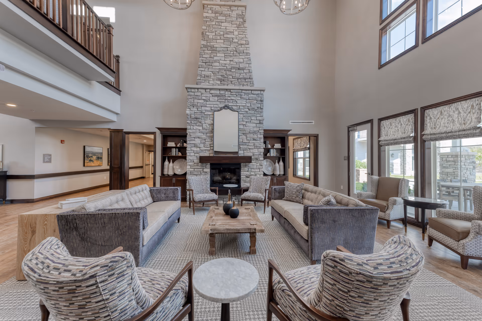 A spacious senior living facility common area with high ceilings and large windows. The room features two long sofas facing each other, two patterned armchairs in the foreground, and additional seating near the windows. A large stone fireplace with a mirror above it is centered on the far wall, flanked by built-in wooden shelves with decorative items. The space is decorated in neutral tones with wooden accents and a light-colored rug covering the floor.