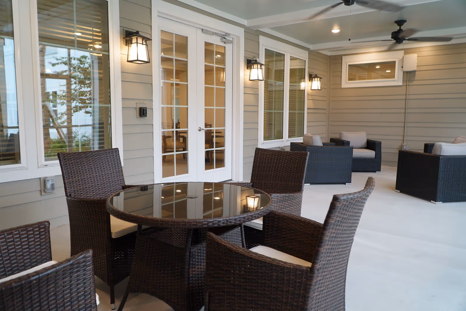 Covered outdoor patio area with a round glass-top table surrounded by four wicker chairs with cushions. In the background, there are additional wicker armchairs with cushions, beige siding walls, white-framed windows and French doors, wall-mounted lantern-style lights, and ceiling fans.