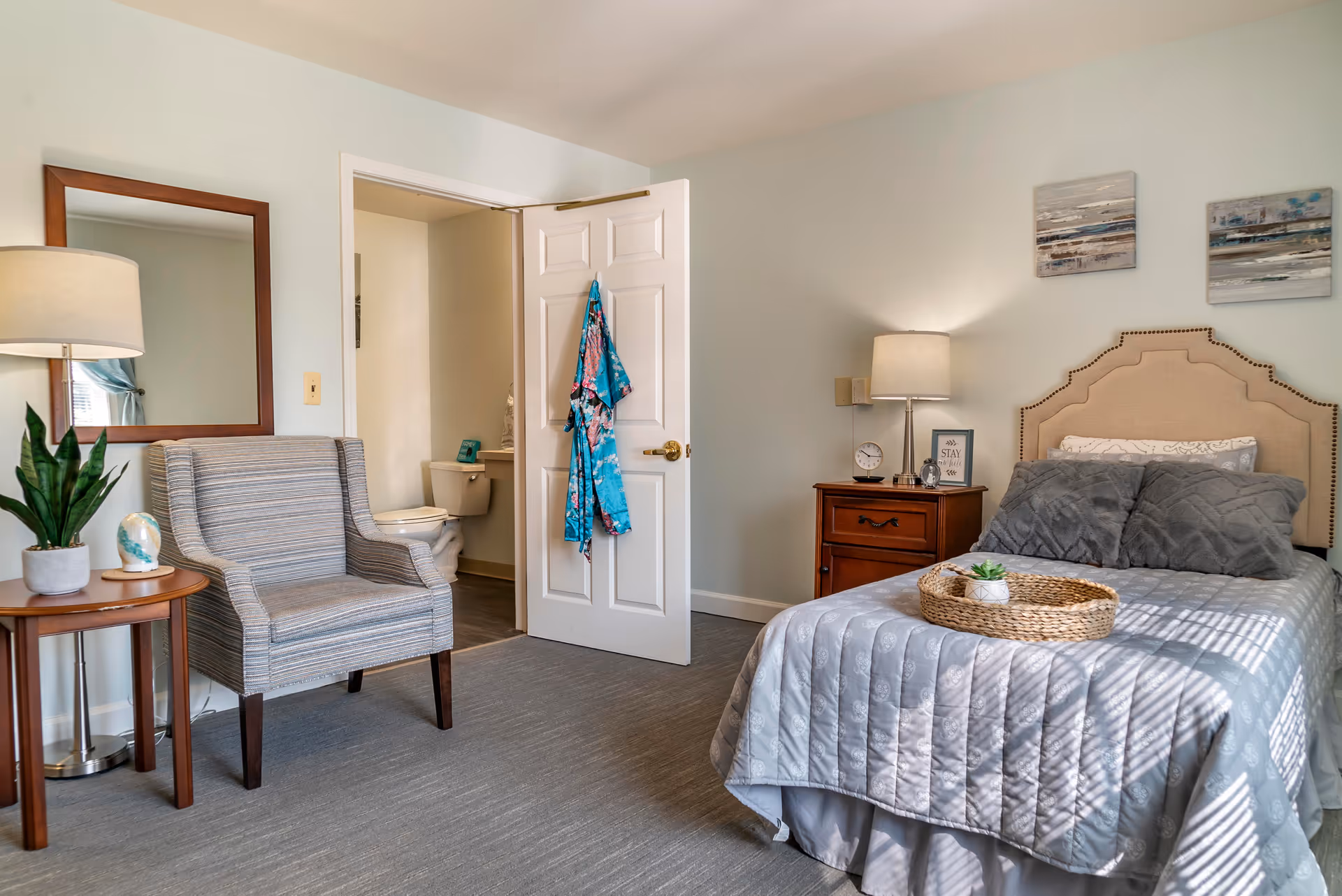 A cozy bedroom with a neatly made bed featuring gray bedding and pillows, a wooden nightstand with a lamp, clock, and framed quote, a striped armchair next to a small round table with a plant and decorative item, a large mirror on the wall, and an open door revealing a bathroom with a toilet. A colorful robe hangs on the door.