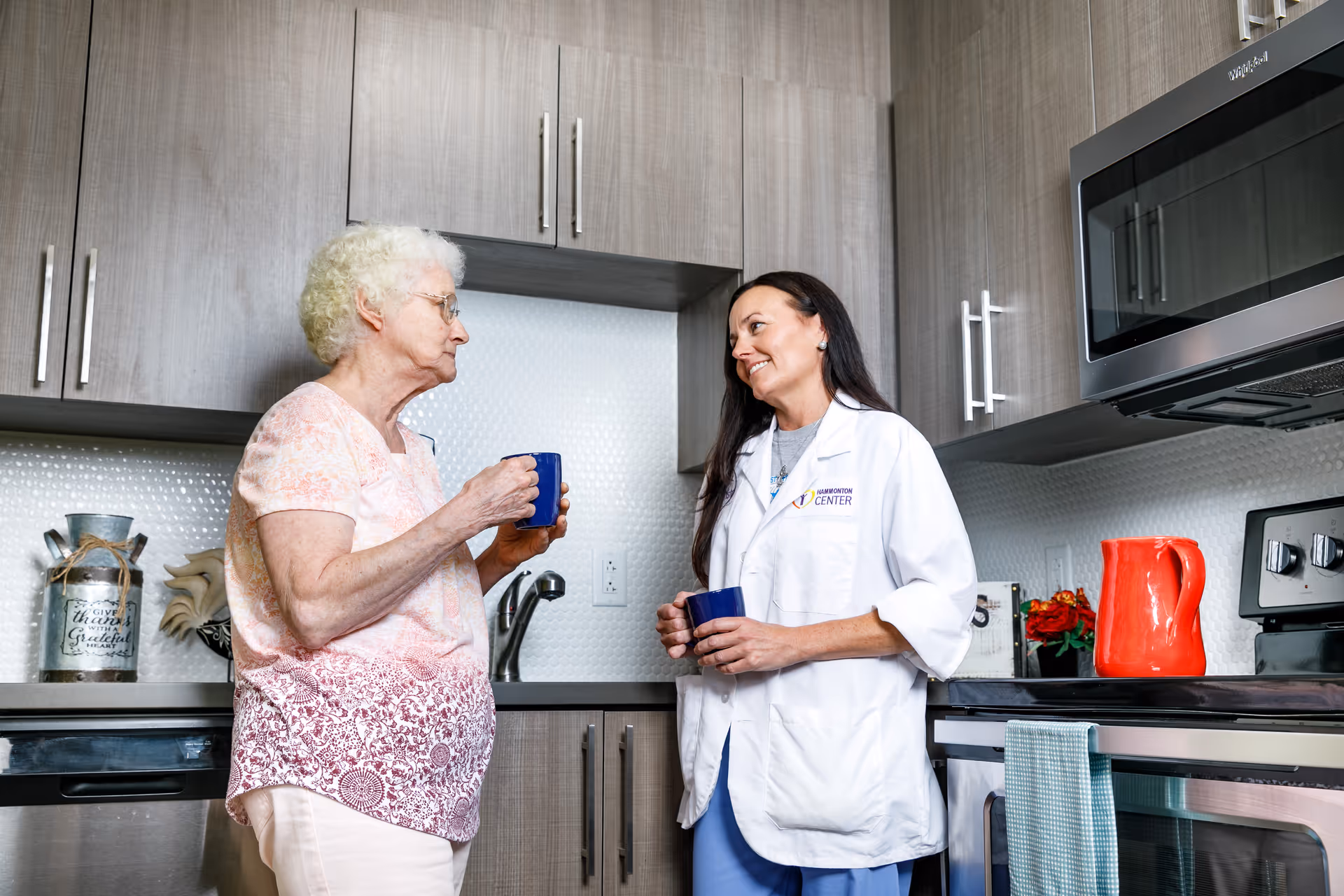 An elderly woman and a healthcare professional in a white coat with Hammonton Center logo are standing in a modern kitchen, holding blue mugs and engaging in a friendly conversation.