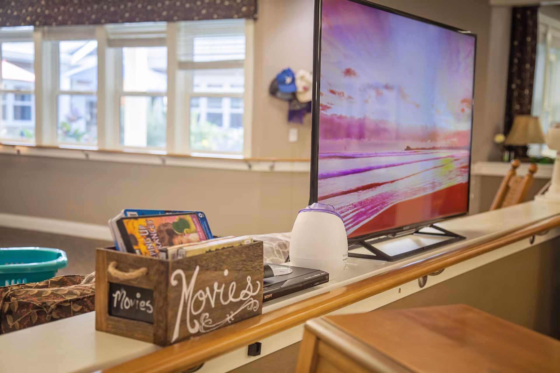 A cozy interior space with a flat-screen TV displaying a colorful beach sunset scene. In front of the TV, there is a wooden box labeled 'Movies' containing DVDs. The room has large windows letting in natural light, a handrail along the wall, and some furniture including chairs and a lamp in the background.