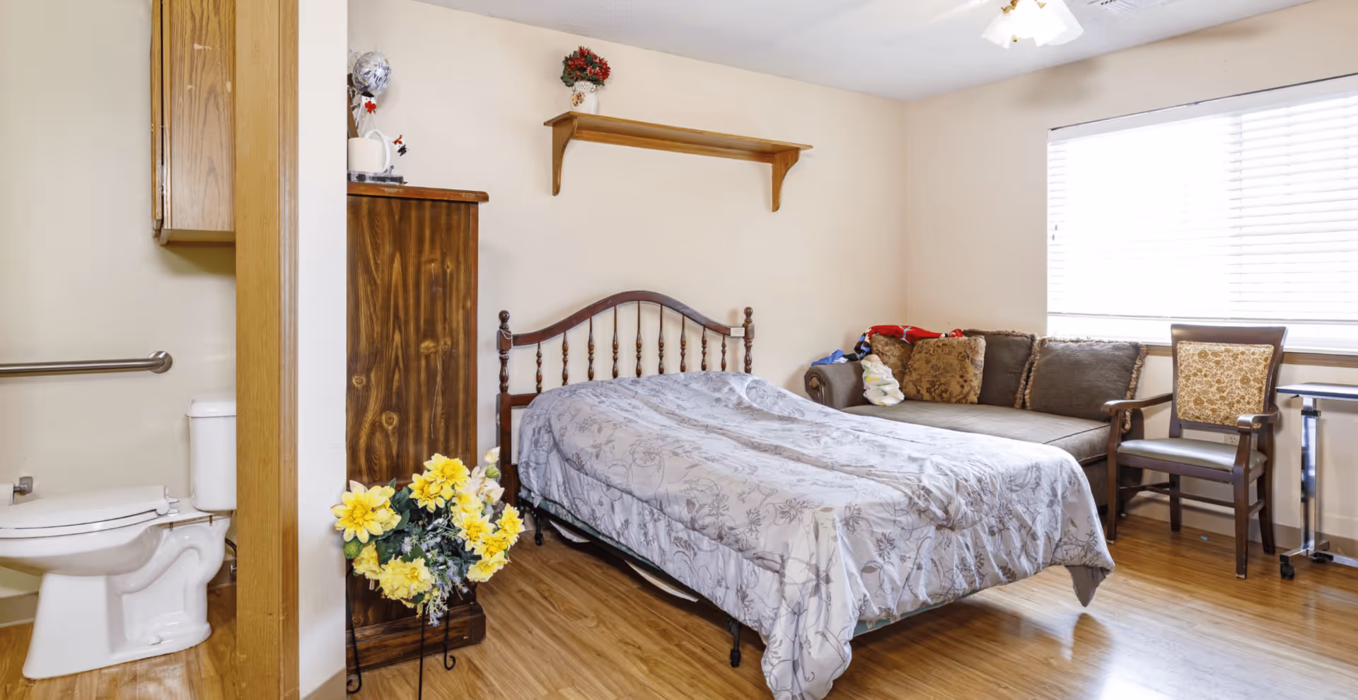 A senior living facility room featuring a bed with a wooden headboard, a brown couch with decorative pillows, a wooden chair, and a window with blinds. To the left, there is a partial view of a bathroom with a toilet and grab bar. The room has wooden flooring and light-colored walls with a small shelf holding a flower vase above the bed.