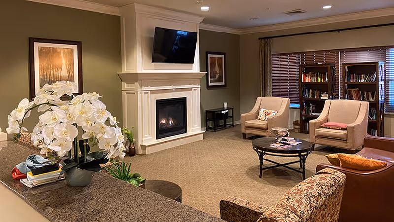 A cozy living room area with a beige carpet, a white fireplace with a TV mounted above it, two beige armchairs with cushions, a round coffee table with magazines, bookshelves filled with books near a window with wooden blinds, and a granite countertop with a vase of white orchids in the foreground.