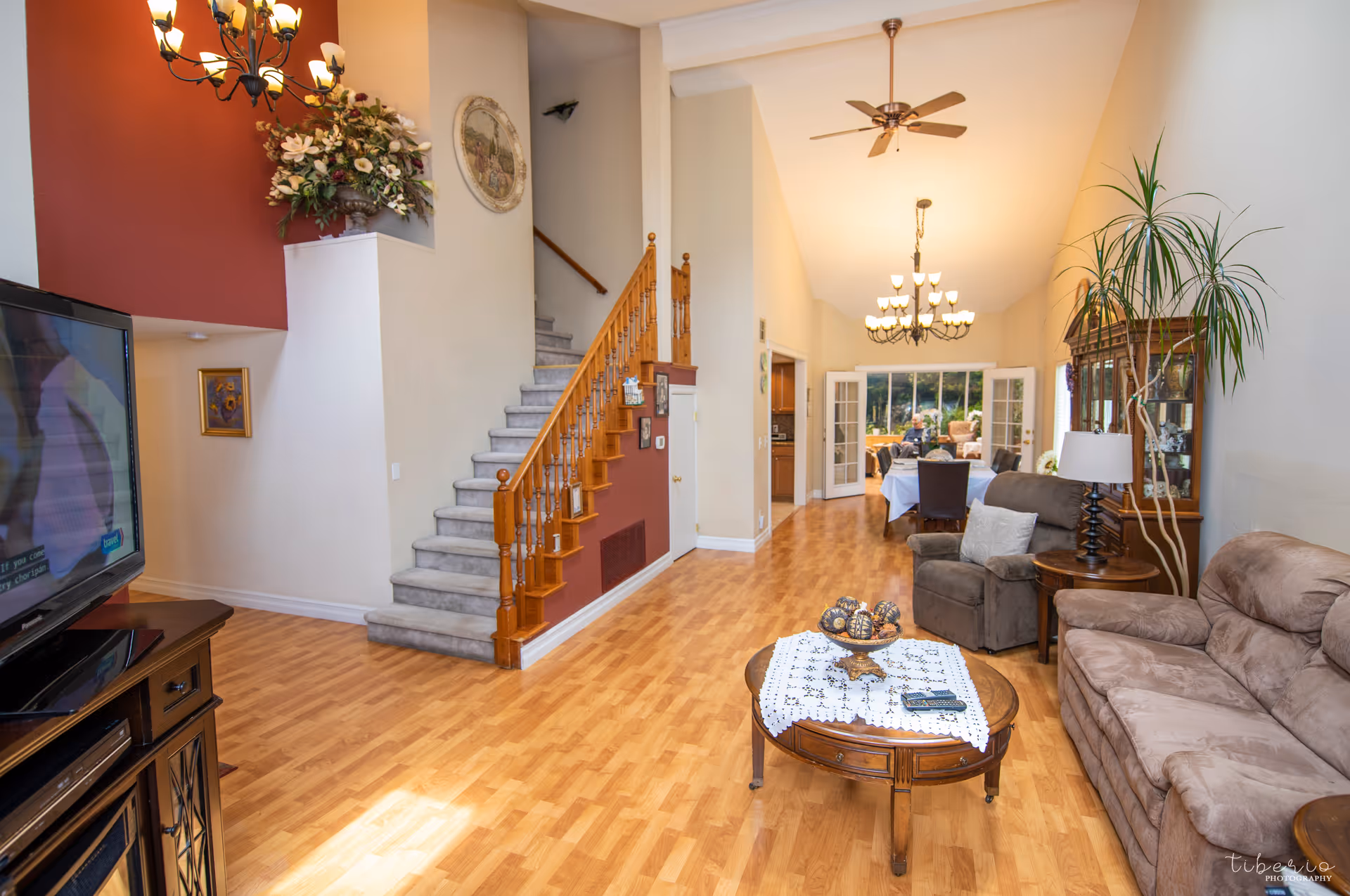 Bright living room with couches, a coffee table, an armchair, a wooden staircase, and a dining area visible in the background.