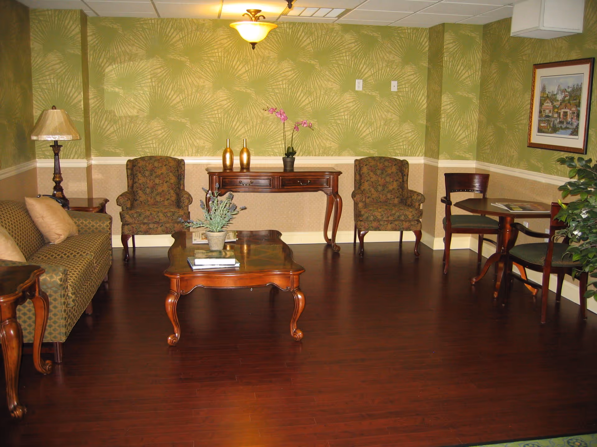 A cozy living room with green patterned wallpaper and dark wood flooring. The room features a green patterned sofa with beige pillows, two floral armchairs, a wooden coffee table with a plant and books, a wooden side table with a lamp, a console table with decorative vases and a potted orchid, and a small round table with two chairs. A framed picture hangs on the wall and there is a ceiling light fixture.