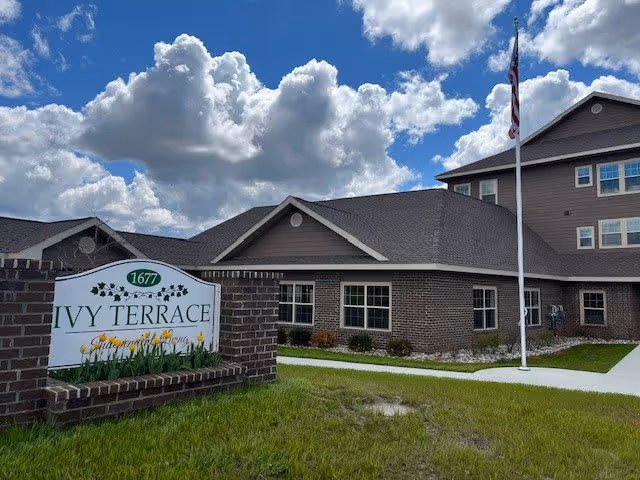 Exterior view of Ivy Terrace of Lapeer independent living facility showing a brick building with multiple windows, a flagpole with an American flag, and a sign in front that reads 'Ivy Terrace 1677'. The sky is partly cloudy with blue patches.