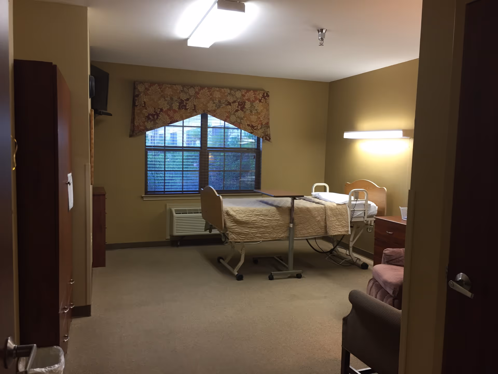 A nursing facility bedroom with a hospital bed covered in a beige quilt, a window with floral valance, an air conditioning unit below the window, a wooden dresser, a pink armchair, and a wall-mounted light above the bed.