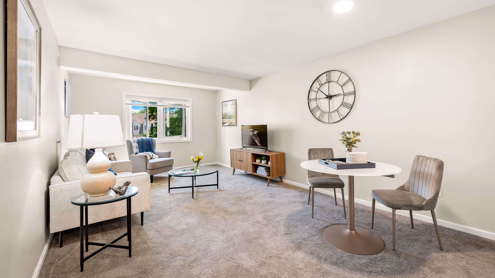 Bright furnished living room with a sofa and armchair, coffee table, TV console, round dining table with two chairs, and a large wall clock.