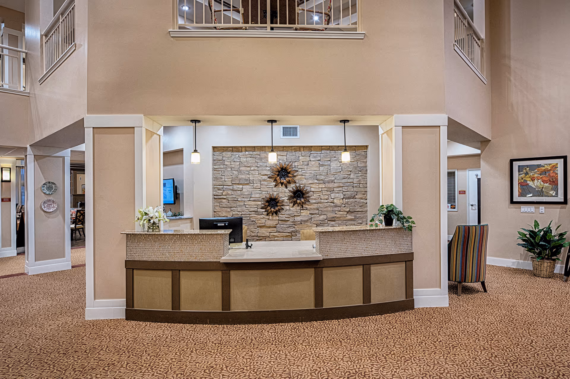 Reception desk area inside The Stonehaven Assisted Living and Memory Care facility with a stone accent wall behind the desk, three hanging pendant lights, decorative plants, and a patterned carpet floor. There is a colorful striped chair and framed artwork on the wall to the right.