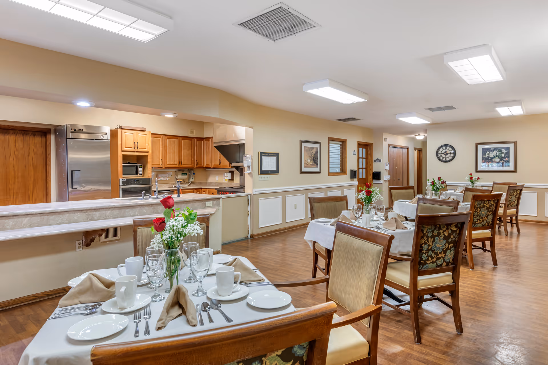 A dining room with several tables set with white tablecloths, plates, cups, silverware, and napkins. Each table has a centerpiece with red roses and white flowers. The room has wooden floors, beige walls with white wainscoting, and framed artwork. In the background, there is a kitchen area with wooden cabinets, a stainless steel refrigerator, and a microwave.