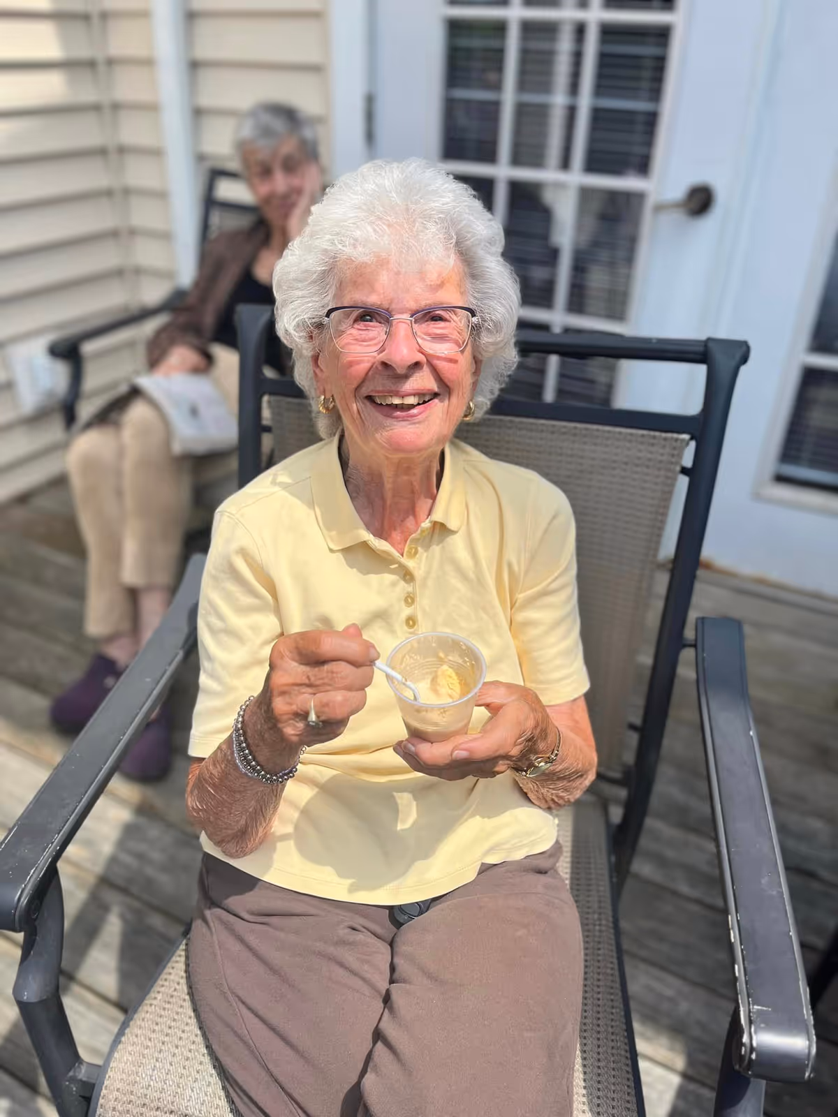 An elderly woman with white hair and glasses is sitting outdoors on a patio chair, smiling and holding a small cup of ice cream with a spoon. Another elderly woman is seated in the background, slightly out of focus, on a similar chair near a house with beige siding and a glass door.