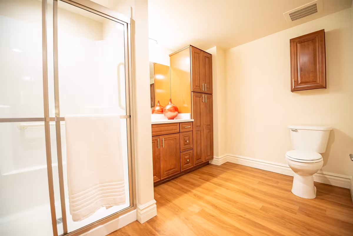 Bright bathroom with a glass-enclosed shower, wooden vanity and cabinets, and a toilet on wood-look flooring.