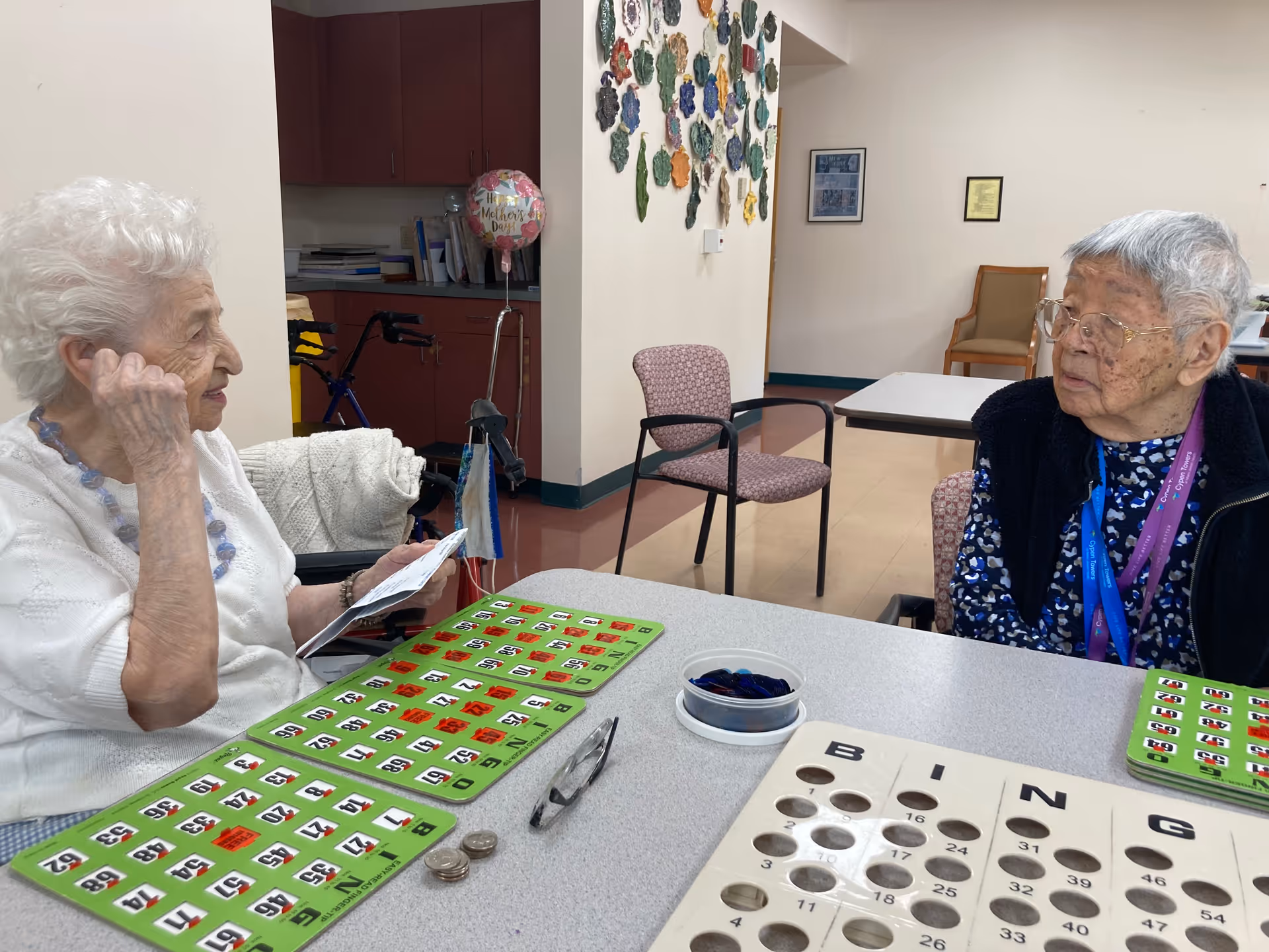 Two elderly women sitting at a table playing bingo in a common area of a senior living facility. One woman is holding bingo cards and appears to be thinking, while the other woman looks on. The table has multiple bingo cards, bingo chips, and some coins. The background shows chairs, a balloon, and wall decorations.