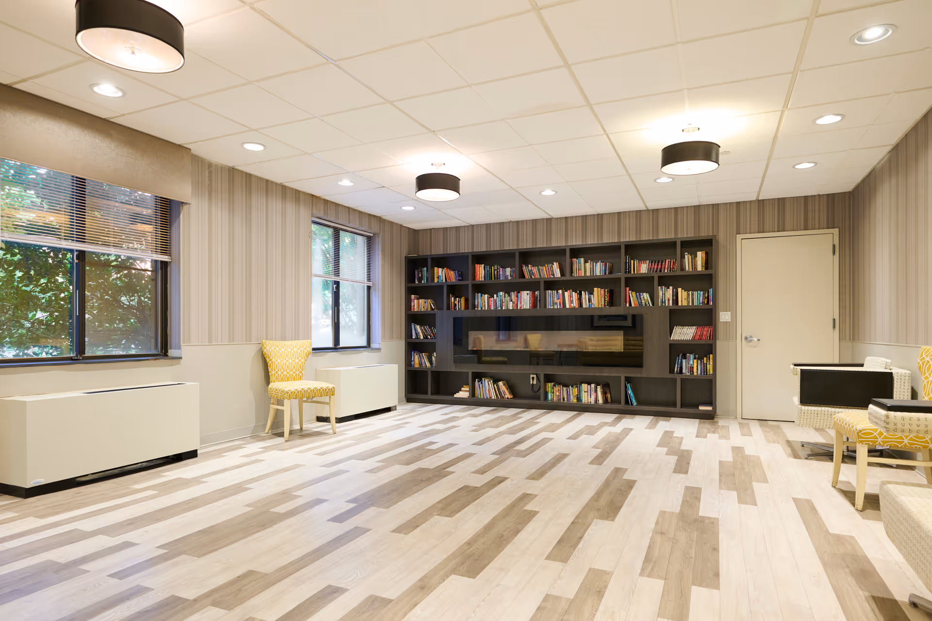 A bright and spacious room with light wood patterned flooring, beige and striped wallpaper, two large windows with blinds, a dark wood bookshelf filled with books, and several yellow patterned chairs placed around the room. The ceiling has recessed lighting and three round black and white light fixtures.