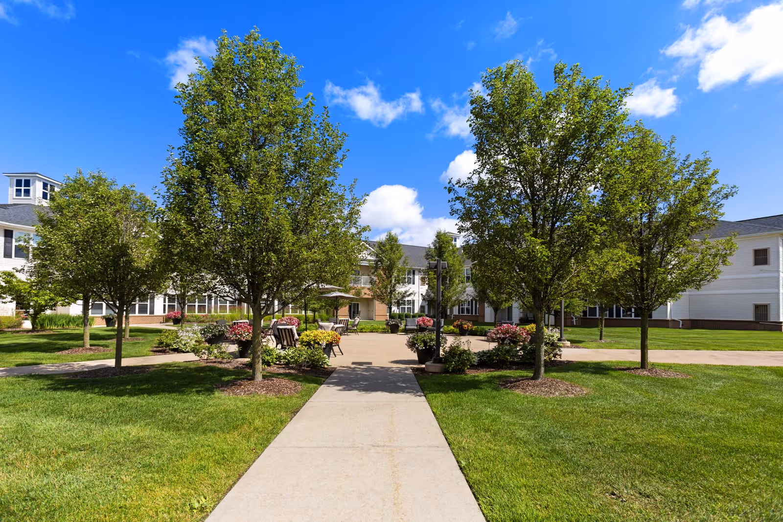 A paved walkway leads through a landscaped garden area with green grass, trees, and flower pots. Benches are placed along the path, and a large building with white siding and multiple windows surrounds the garden under a bright blue sky with scattered clouds.