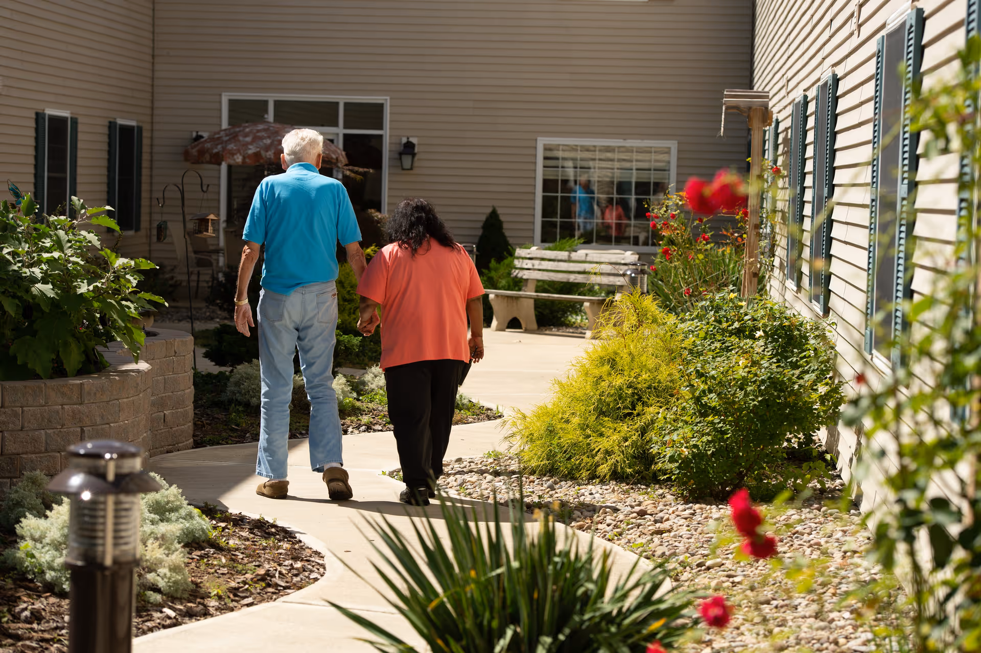 An elderly man and woman walking hand in hand along a curved garden pathway outside a beige building with windows and green shutters. The garden is landscaped with various plants, flowers, and shrubs, and there is a bench and patio umbrella in the background.