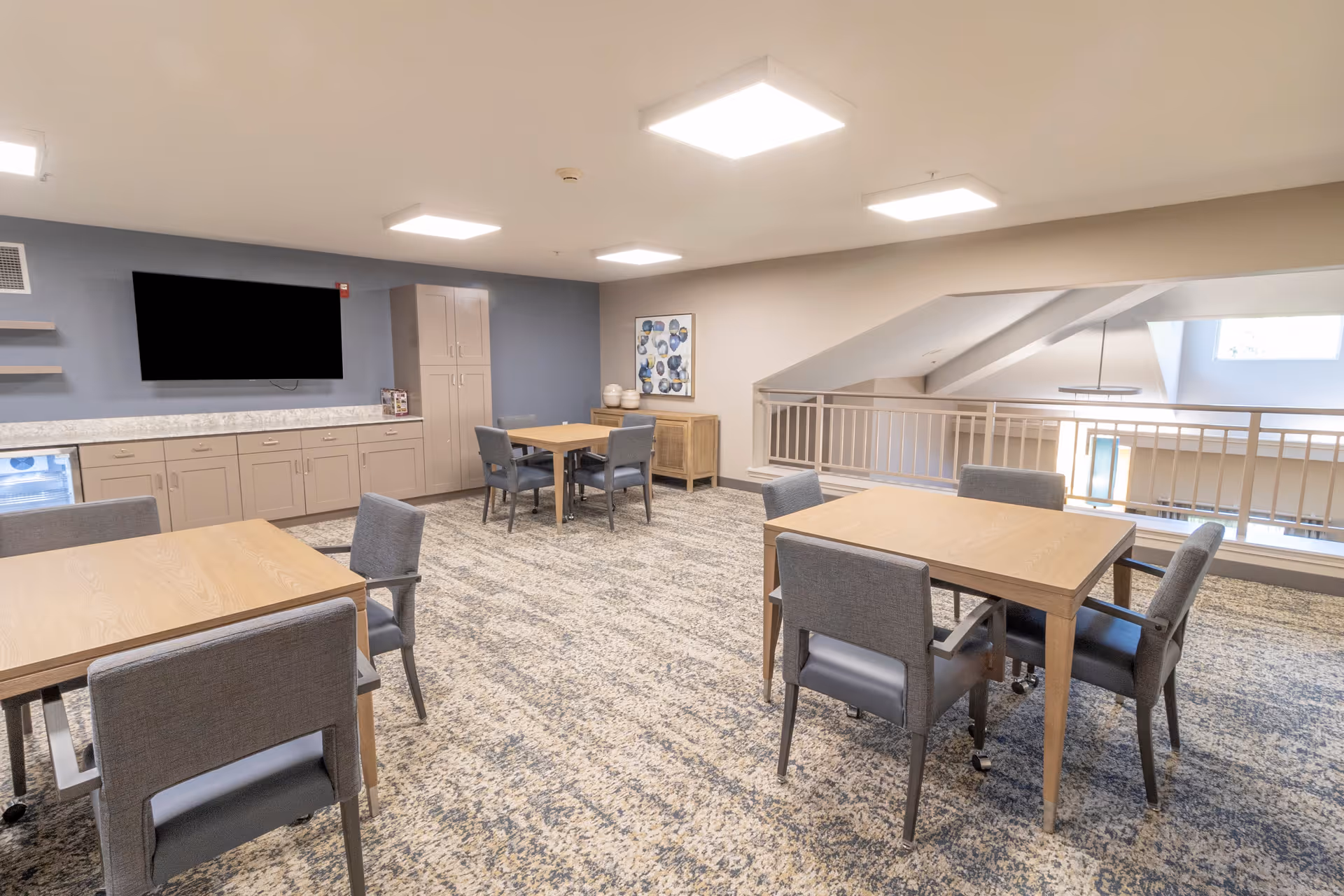 A bright and spacious common area with several wooden tables and gray upholstered chairs arranged around them. The room features a carpeted floor, a large flat-screen TV mounted on a blue-gray wall, and built-in cabinetry with a countertop. There is a railing overlooking a lower level, and the ceiling has recessed lighting panels.