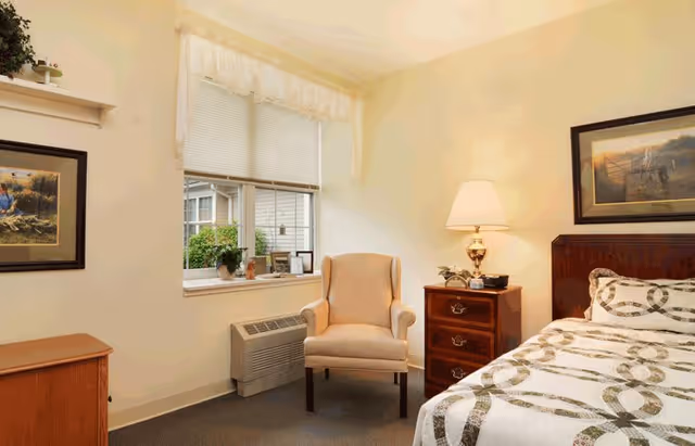 A cozy bedroom with a single bed featuring a patterned quilt, a wooden nightstand with a lamp, a beige armchair, a window with blinds and a valance, and framed artwork on the walls.