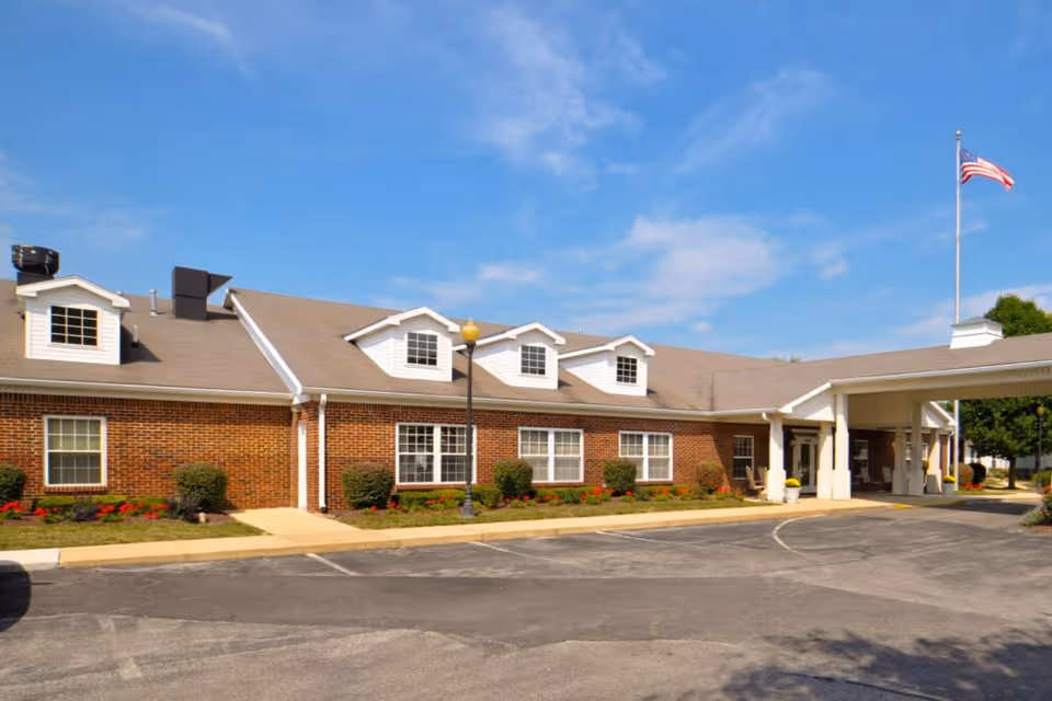 Exterior view of a single-story brick building with multiple windows, a covered entrance, and an American flag flying on a flagpole under a blue sky with some clouds. There are small bushes and flowers planted along the building and a paved driveway in front.