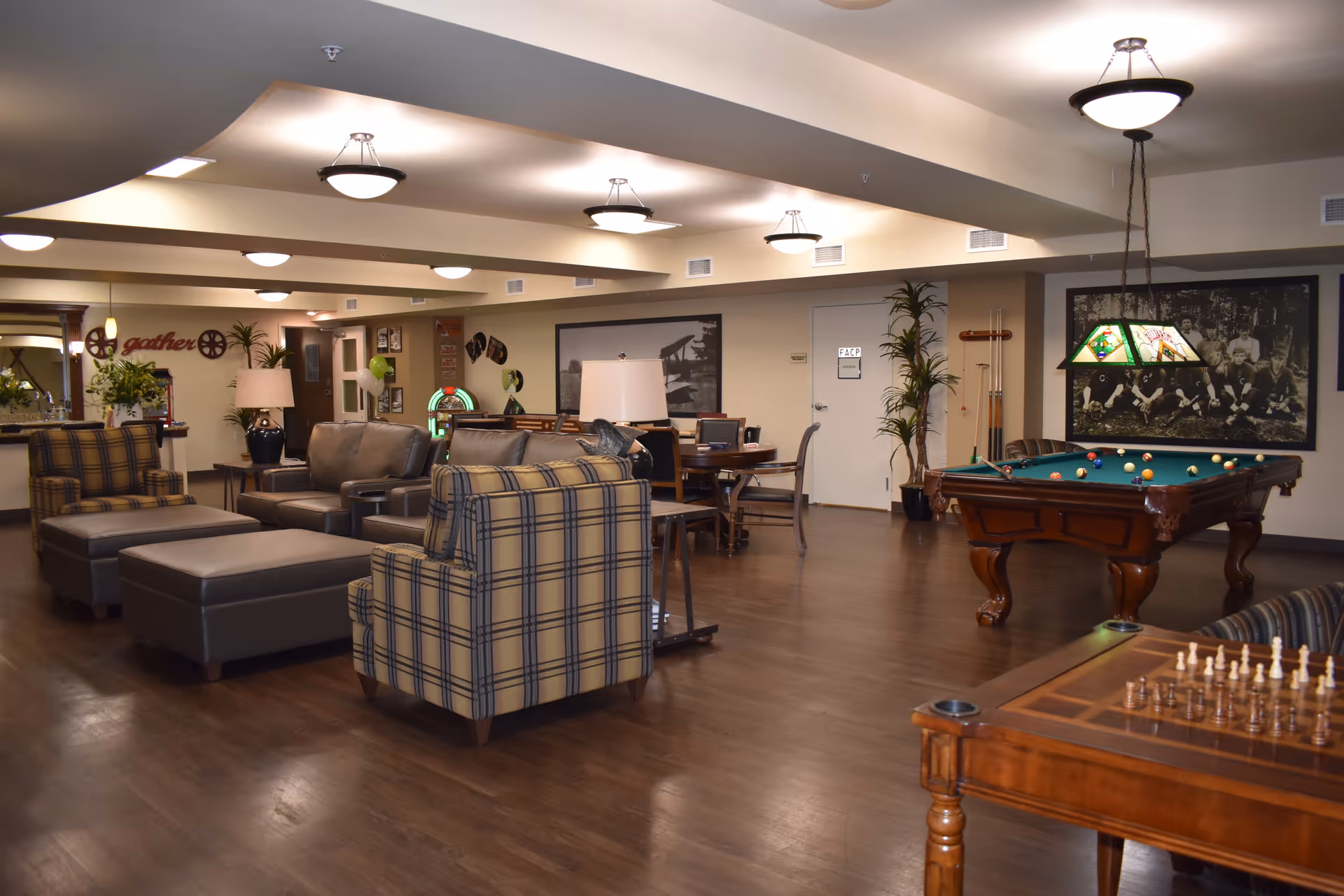 A spacious senior living facility common area with comfortable seating including plaid and leather armchairs, a pool table with balls set up, a chess table, and a dining table in the background. The room is well-lit with ceiling lights and decorated with plants and framed pictures on the walls.
