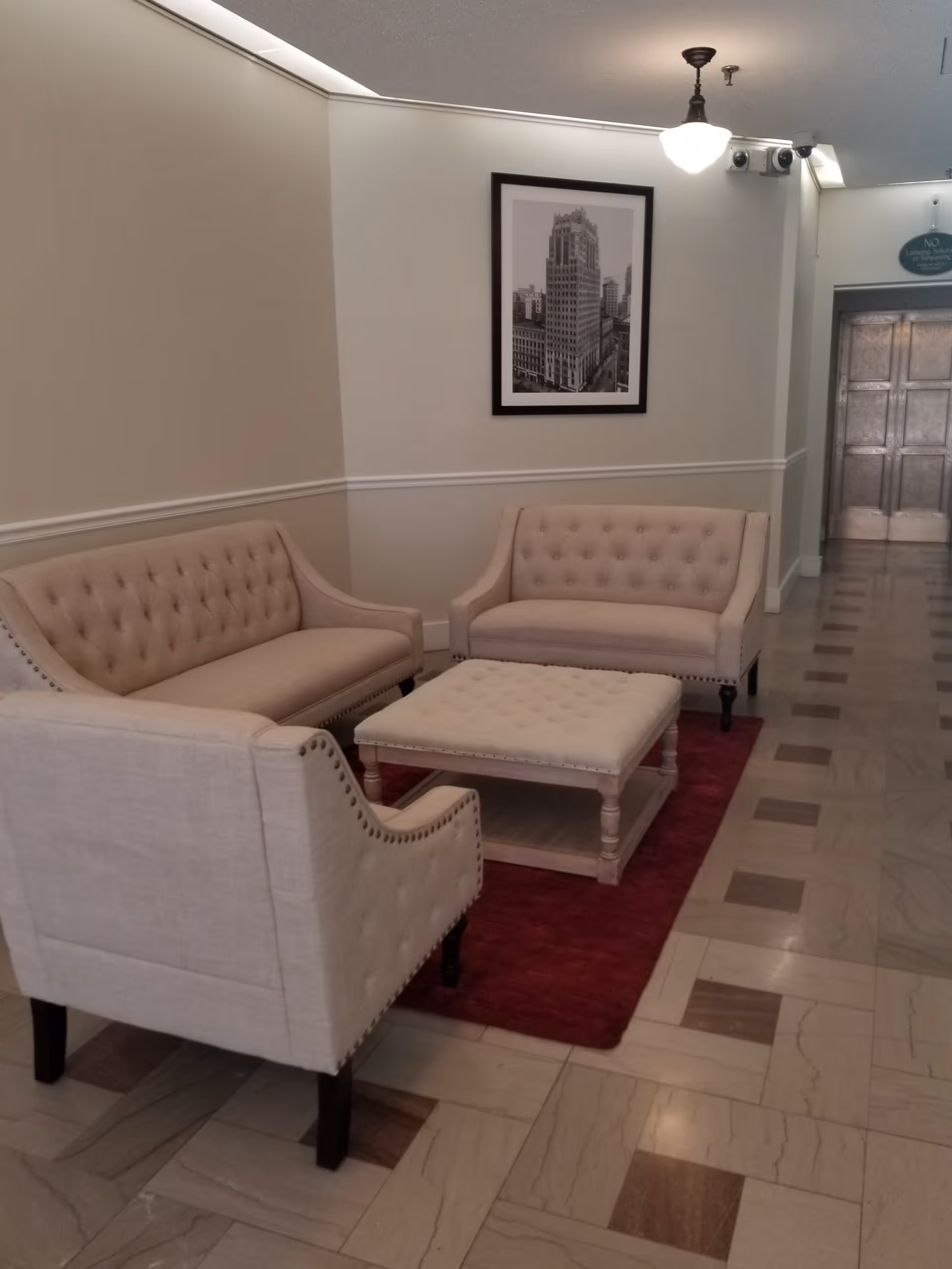 A seating area in a hallway with two beige tufted loveseats, a matching armchair, and a tufted ottoman on a red rug. A black and white framed photograph of a tall building hangs on the wall above the loveseats. The floor is tiled with a beige and brown pattern, and a light fixture hangs from the ceiling.