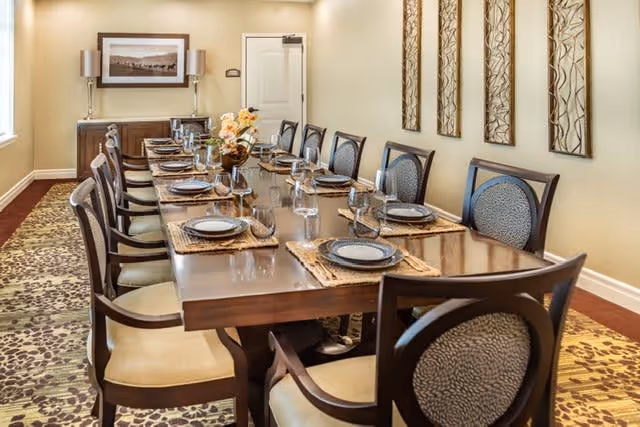 A long wooden dining table set for twelve people with plates, glasses, and silverware in a well-lit dining room. The room has beige walls adorned with three decorative wall panels and a framed picture above a sideboard. The floor is covered with a patterned carpet.