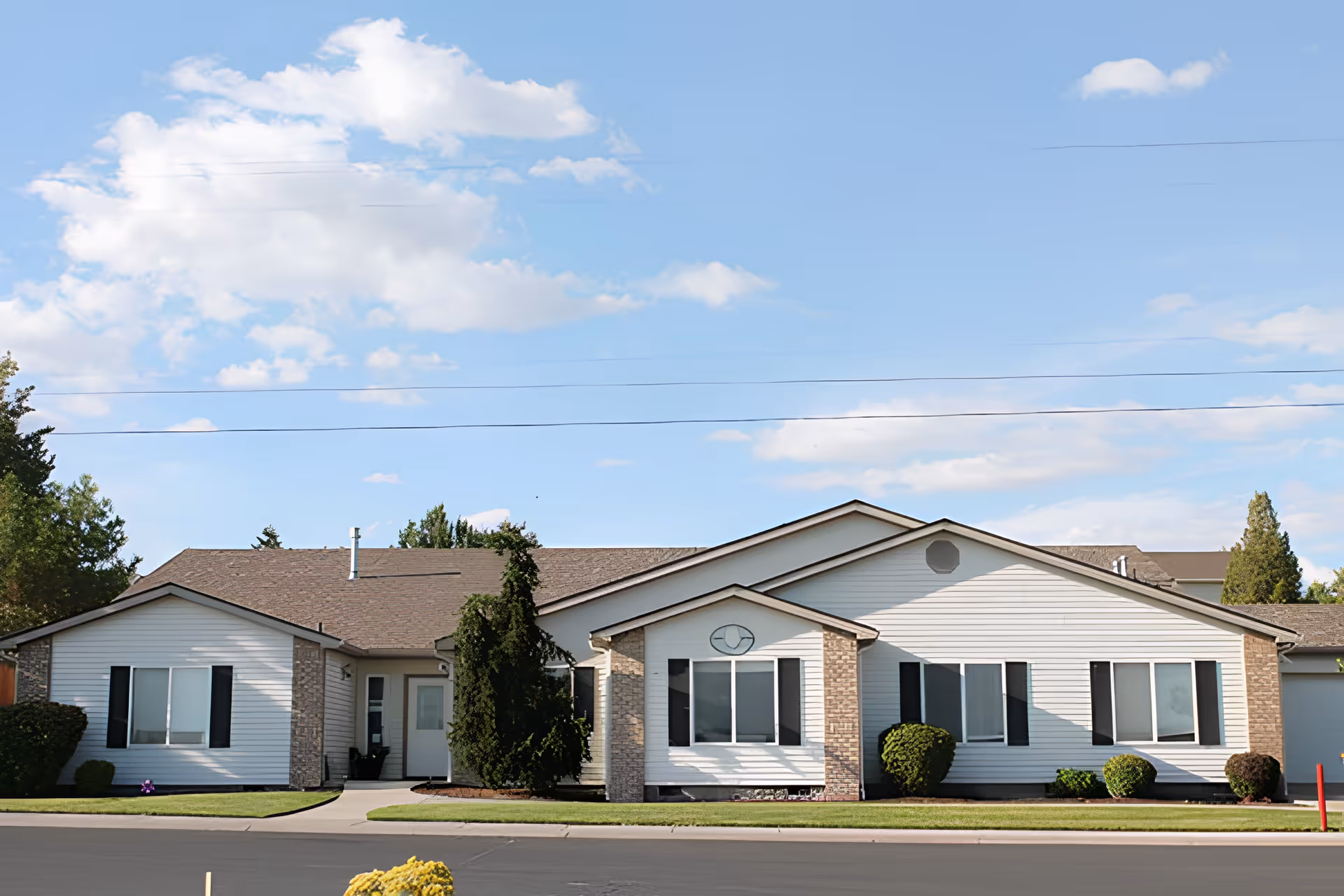 Front view of a single-story white siding senior living building with a manicured lawn and shrubs under a blue sky.