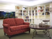 A cozy living room area featuring a red patterned loveseat, a round wooden coffee table with ornate metal legs, and built-in white bookshelves filled with books and decorative items. There is also a large fish tank integrated into the shelving unit.