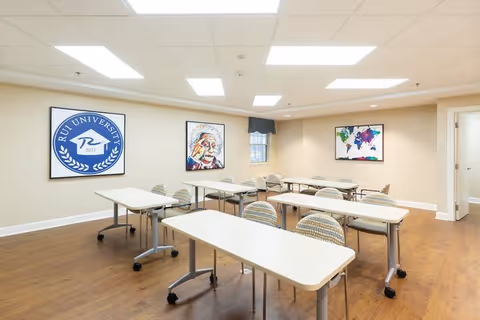 A well-lit classroom or meeting room with several white tables and striped chairs arranged in rows on a wooden floor. The walls are beige and decorated with three framed artworks, including a RUI University logo, a portrait of Albert Einstein, and a colorful world map. The ceiling has recessed lighting panels.