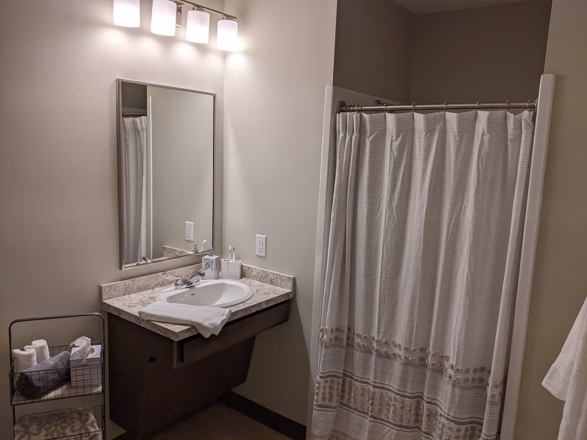 Bathroom with a sink and mirror under a three-light fixture, a towel rack with folded towels, and a shower with a patterned curtain.
