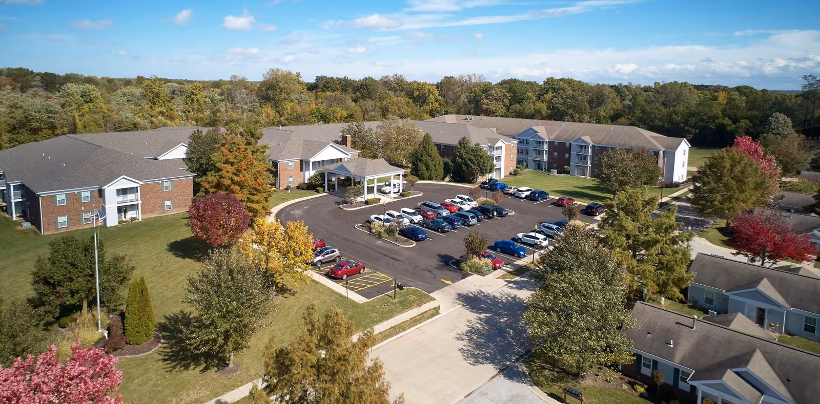 Aerial view of Asbury Village senior living facility showing multiple brick and siding buildings surrounded by trees with autumn foliage, a parking lot with several cars, and well-maintained lawns and landscaping under a partly cloudy blue sky.