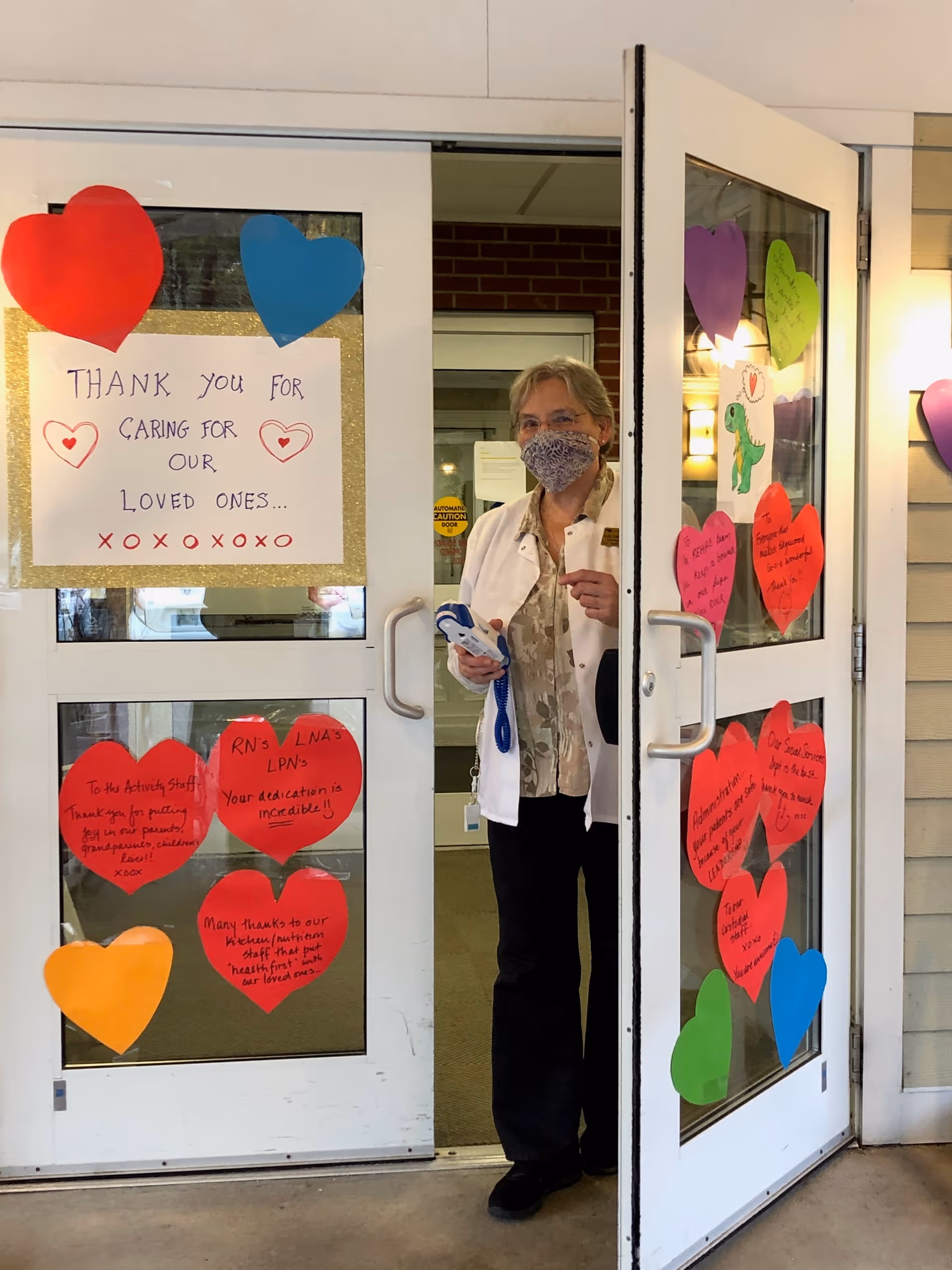 A woman wearing a face mask and white coat stands in the doorway of a building decorated with colorful paper hearts and a sign that reads 'Thank you for caring for our loved ones... xoxoxoxo'. The hearts contain handwritten messages of gratitude.