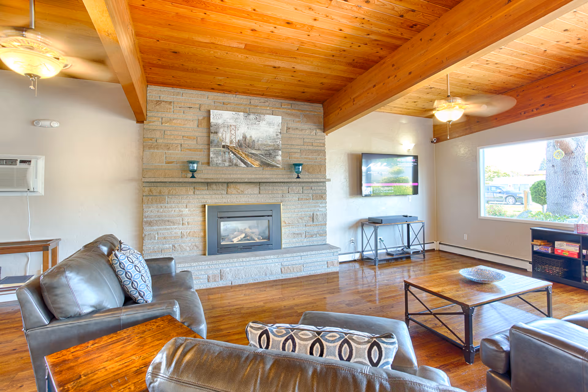 A cozy living room with wooden ceiling beams and a stone fireplace. The room features leather sofas with patterned cushions, a wooden coffee table, a wall-mounted TV, and a large window letting in natural light. Ceiling fans with lights are mounted on the wooden ceiling.