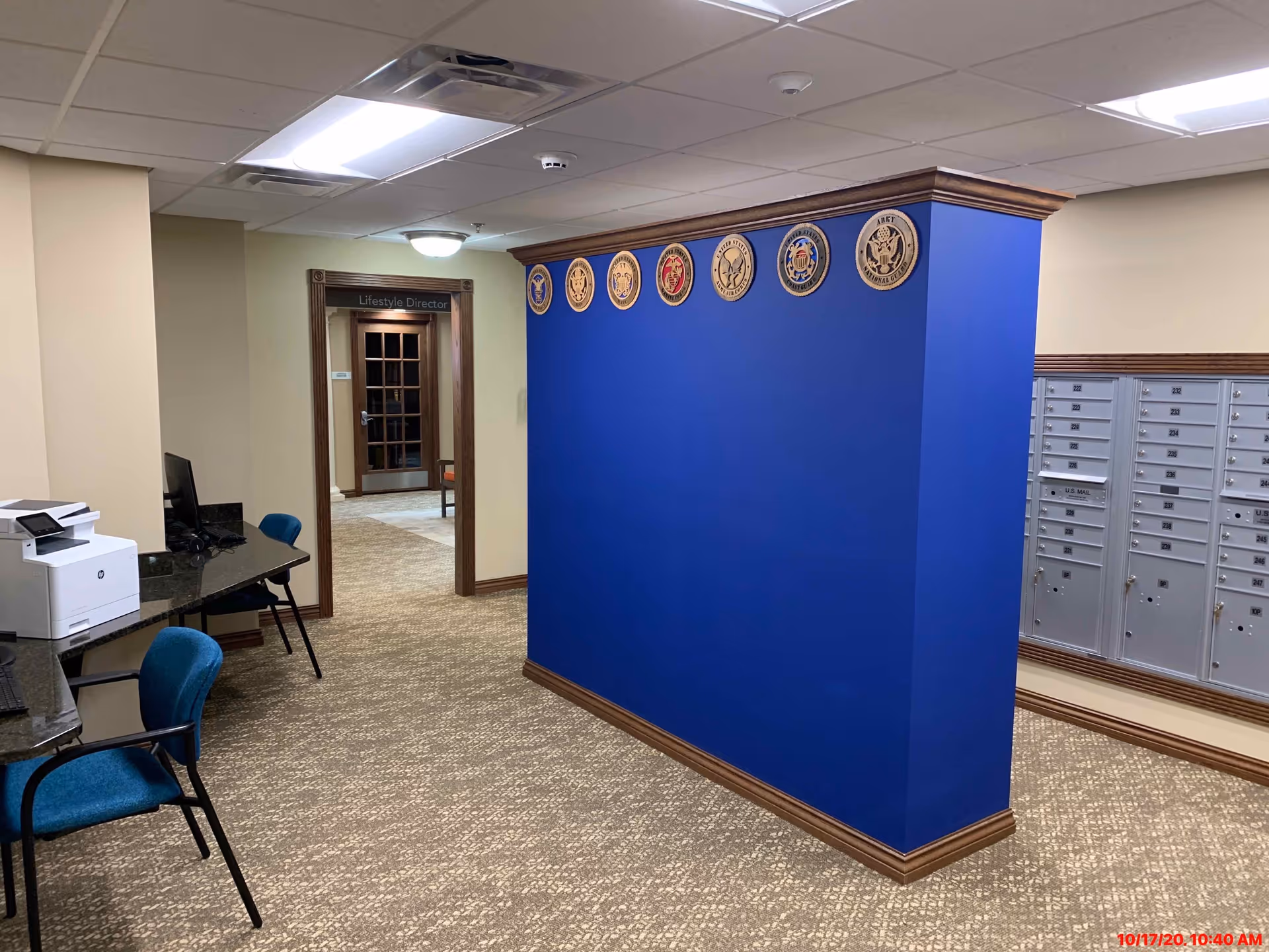 Interior hallway of a senior living facility with a blue partition wall displaying military emblems along the top. To the right are multiple mailboxes, and to the left is a small office area with a printer and chairs. A doorway labeled 'Lifestyle Director' is visible in the background.