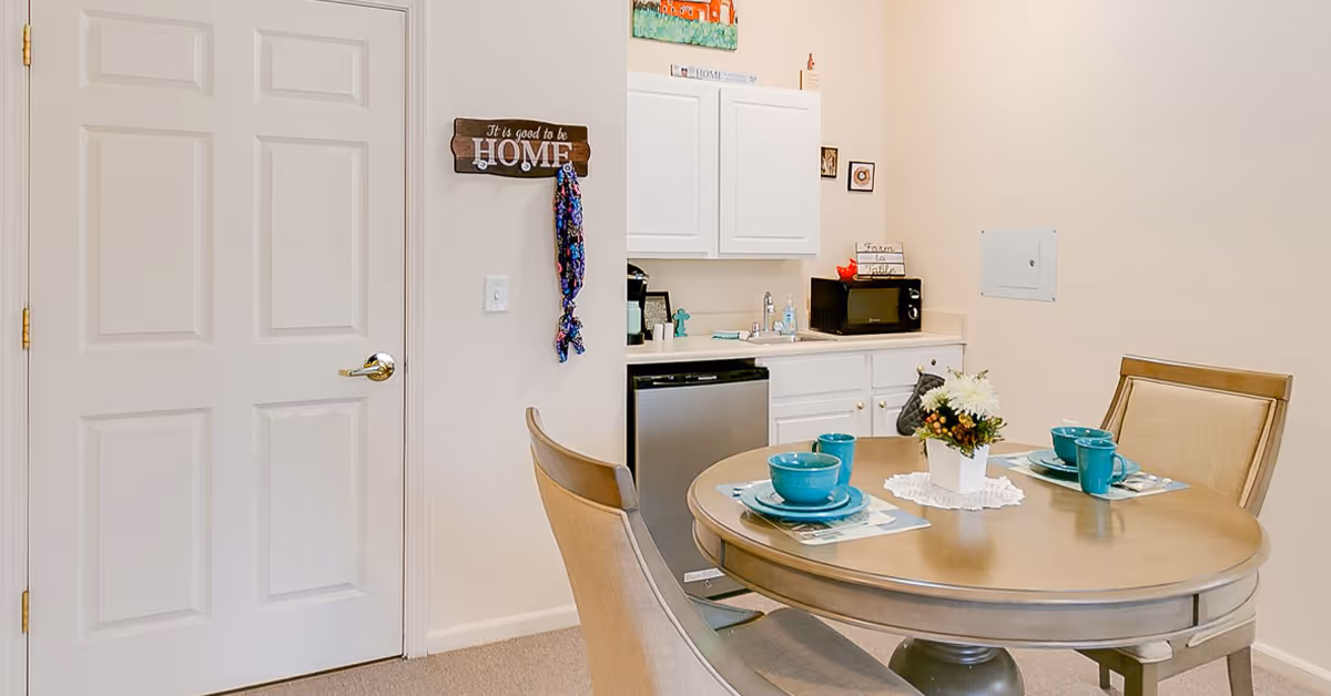 A small dining area with a round wooden table set for two with teal bowls, plates, and mugs. There are two beige upholstered chairs around the table. In the background, there is a compact kitchenette with white cabinets, a small sink, a microwave, and a mini refrigerator. A decorative sign on the wall reads 'It is good to be HOME' with a colorful scarf hanging from it.