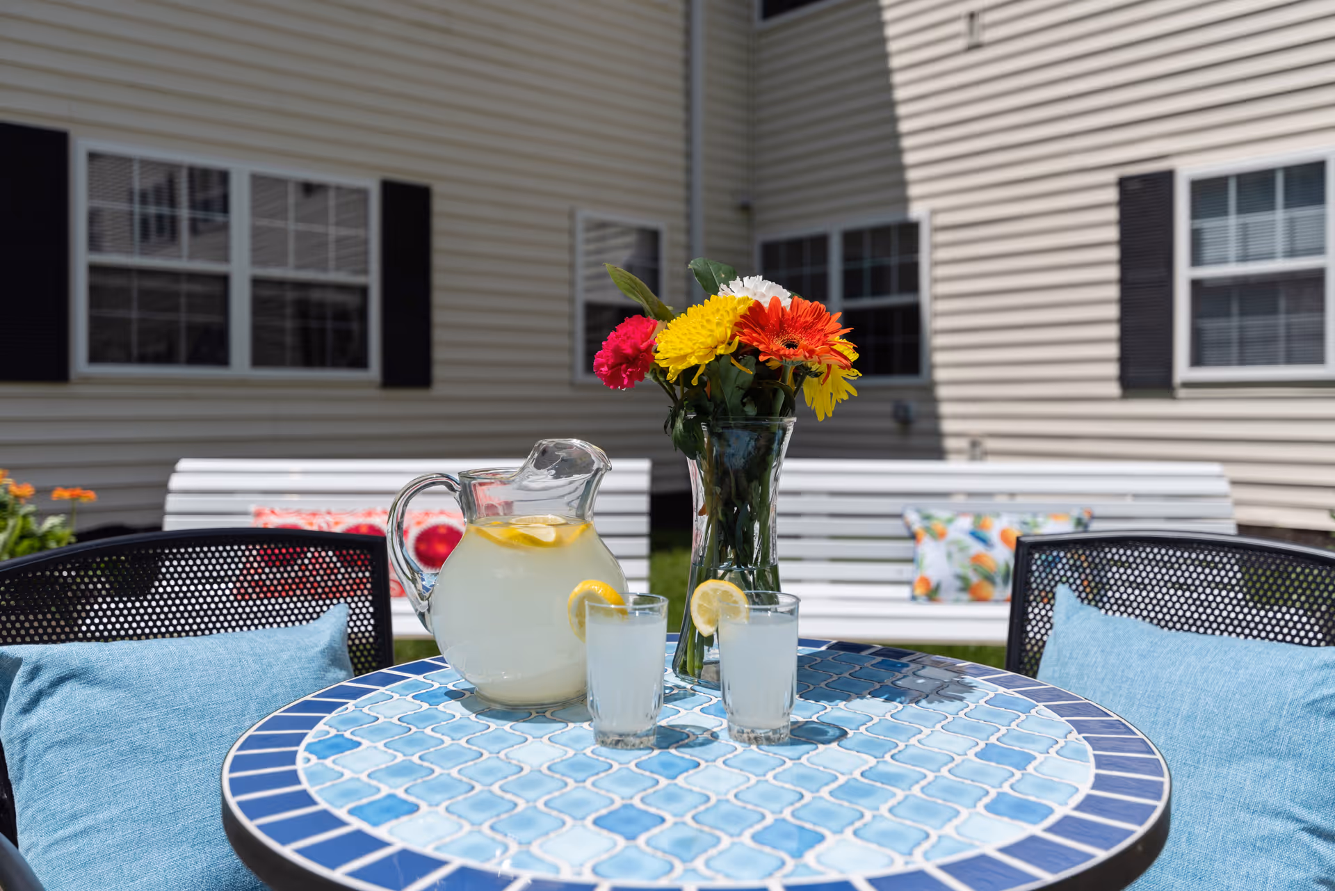 Outdoor patio area with a round blue mosaic table holding a pitcher of lemonade, two glasses with lemon slices, and a vase of colorful flowers. In the background, there are white benches with decorative pillows against the beige siding of a building with windows.