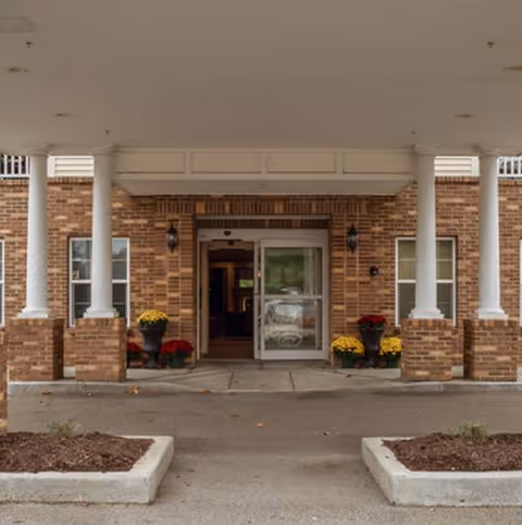 Brick senior living facility entrance with columns, glass doors, and colorful potted flowers.