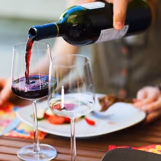 A close-up of a hand pouring red wine from a bottle into a wine glass on a dining table, with another empty wine glass and a plate of food in the background.