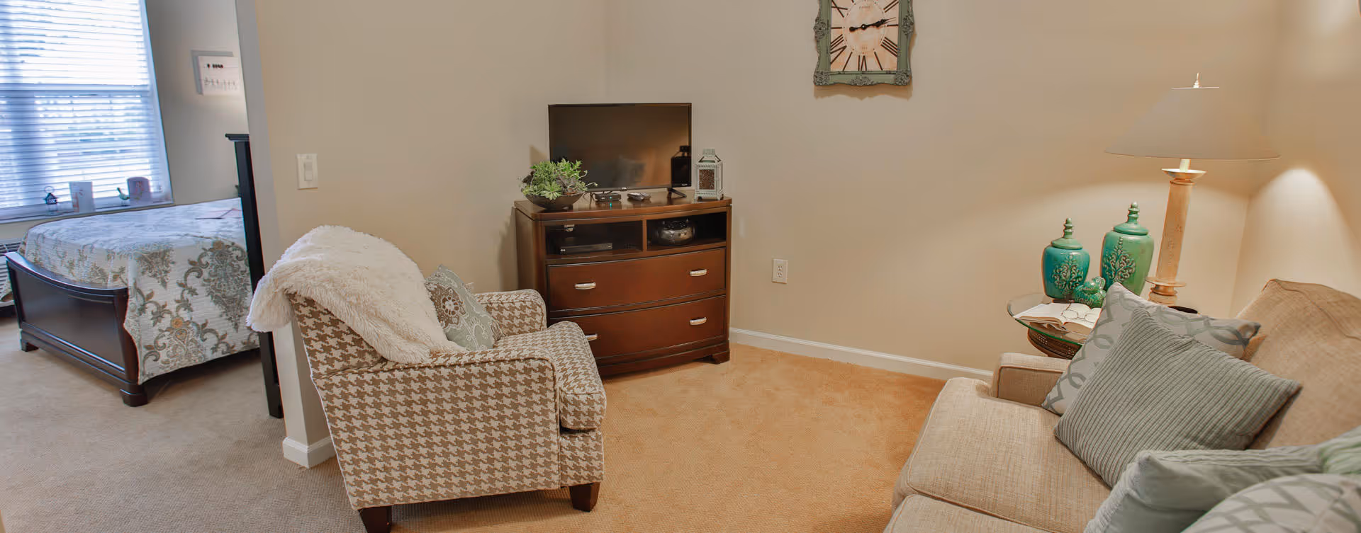 A cozy living area with a beige sofa adorned with multiple cushions, a patterned armchair with a throw blanket, a wooden TV stand with a flat-screen TV and decorative items, a side table with a lamp and green vases, and a wall clock. In the background, a bedroom with a bed covered in a floral quilt is partially visible through an open doorway.